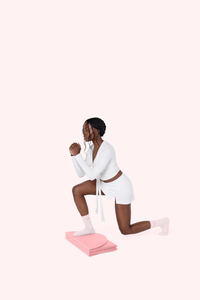 Woman in white outfit exercising on a pink Bala Compact mat against a white background