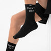 A person wearing black crew socks with white text reading 'CHEAT DAY' on the upper cuff, shown against a clean white background.