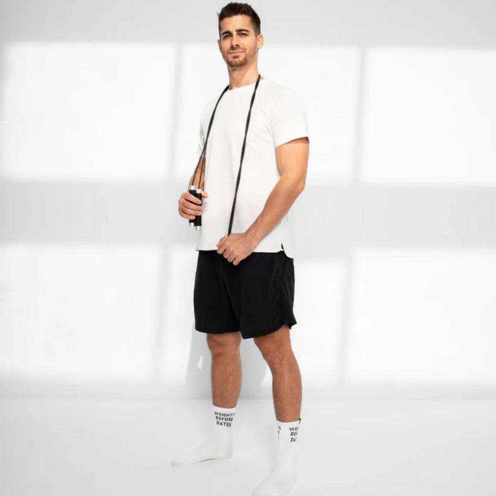 A man modeling a white crew-neck t-shirt and black athletic shorts, paired with white crew socks, standing in a bright, minimalist studio with soft window-like shadows.