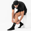 A man in a black t-shirt and shorts sits on a white block, adjusting a black crew sock that has the text 'CHOOSE DAY' printed in white on the cuff.
