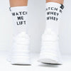 White crew socks with black text reading 'WATCH ME LIFT' worn with chunky white sneakers, shown from the back on a person's feet against a white background.
