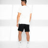 A man shown from behind wearing a white t-shirt, black athletic shorts, and white socks, standing against a bright white studio background.