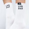 White crew socks worn on feet, featuring the words 'LEG DAY' in black block letters on the upper leg and a small black brand tag near the cuff that reads 'GYM SOX'.