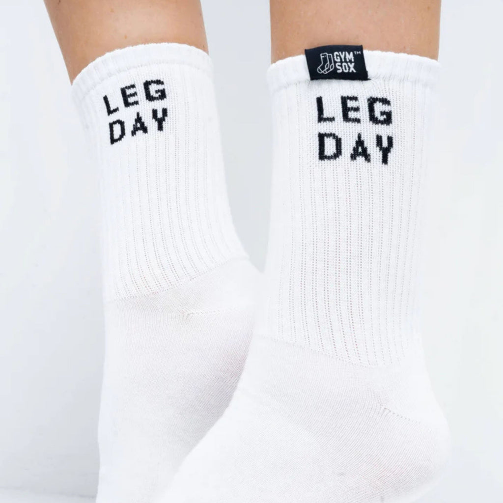 White crew socks worn on feet, featuring the words 'LEG DAY' in black block letters on the upper leg and a small black brand tag near the cuff that reads 'GYM SOX'.