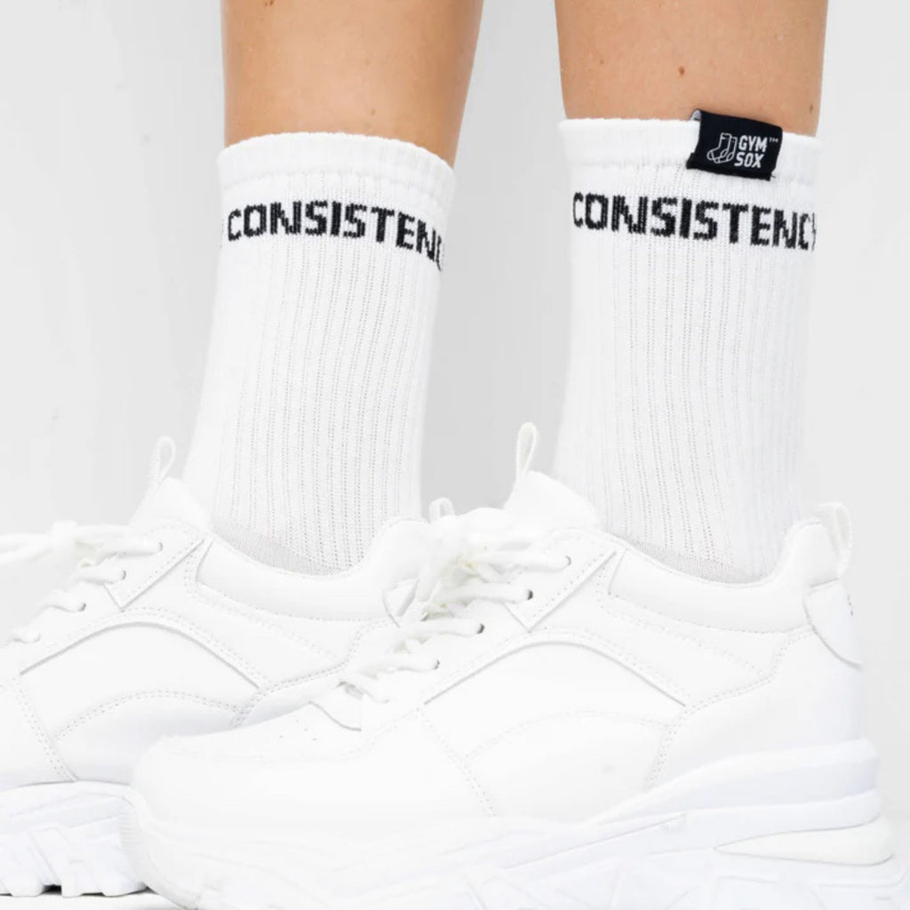 White socks with 'CONSISTENT' text worn with white sneakers on a light gray background