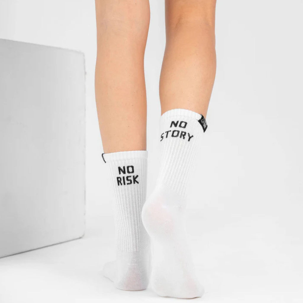 White crew socks worn on a person's feet, featuring black text that reads 'NO RISK' on one sock and 'NO STORY' on the other, photographed against a clean white studio background.
