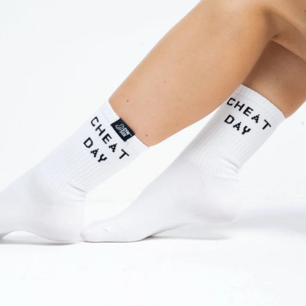 White crew socks worn on a person's feet, featuring the text 'CHEAT DAY' printed in black on the sides and a small black brand tag near the cuff.