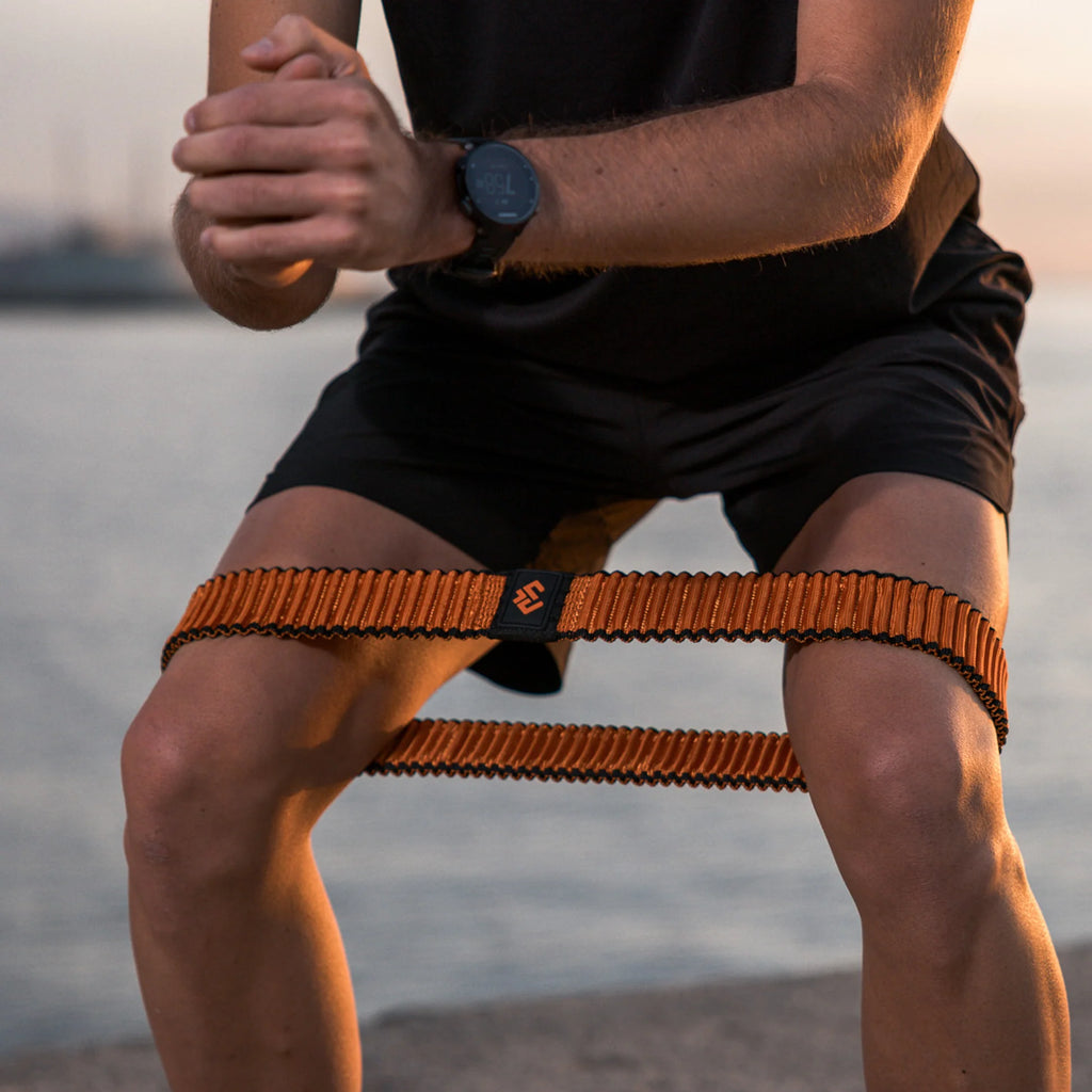 Person sitting on a beach with an orange resistance band, wearing a black watch.