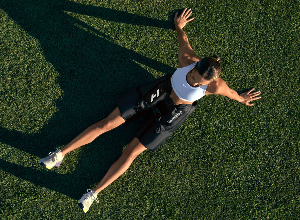 Black compression hip recovery tool by Hyperice worn on a woman lying on a grass surface with her arms outstretched.
