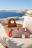 A styled collection of travel and beach accessories arranged on a beige outdoor lounge chair overlooking a blue sea and white buildings. The set includes a woven straw tote with a brown leather handle, a dusty pink pouch with a red strap and clasp, a clear zip pouch with a pink border containing pink items, and a small clear bottle with a pink cap.