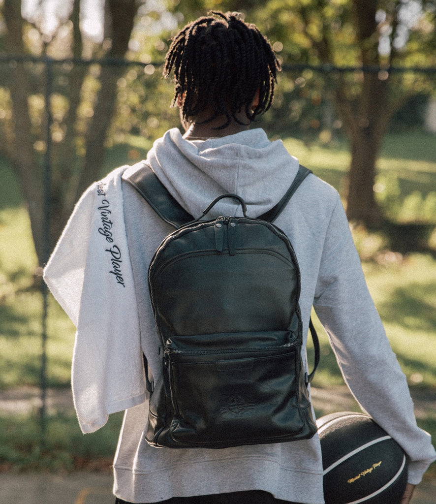 Back side of a person wearing a brown leather backpack with a front pocket and visible stitching, and holding a basketball, displayed against a tree landscape.
