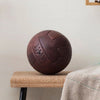 A brown, vintage-style T-panel soccer ball with embossed text and logo on a bench in front of an off-white wall.