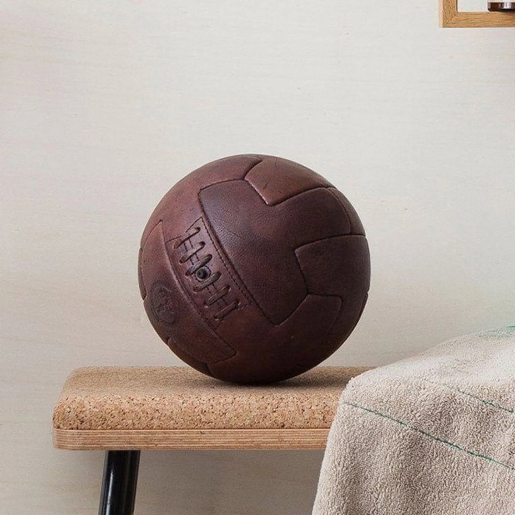 A brown, vintage-style T-panel soccer ball with embossed text and logo on a bench in front of an off-white wall.