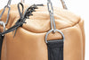 Close-up of a tan leather bag with black straps and metal hardware on a white background