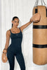 Person in a navy blue athletic outfit with boxing gloves holding a beige punching bag against a white curtain background.