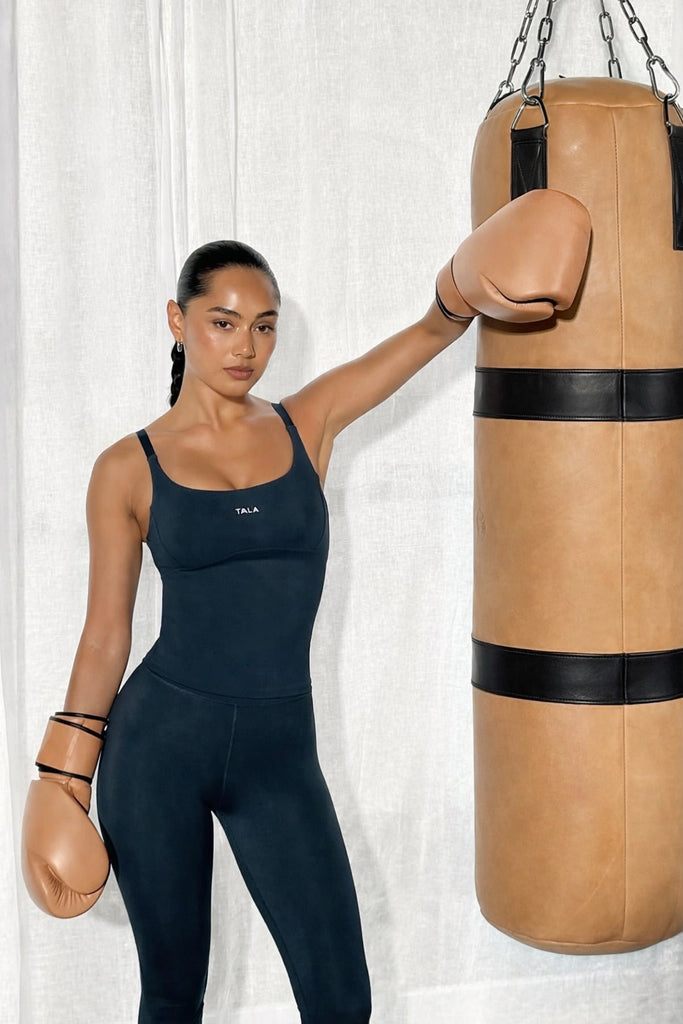 Person in a navy blue athletic outfit with boxing gloves holding a beige punching bag against a white curtain background.